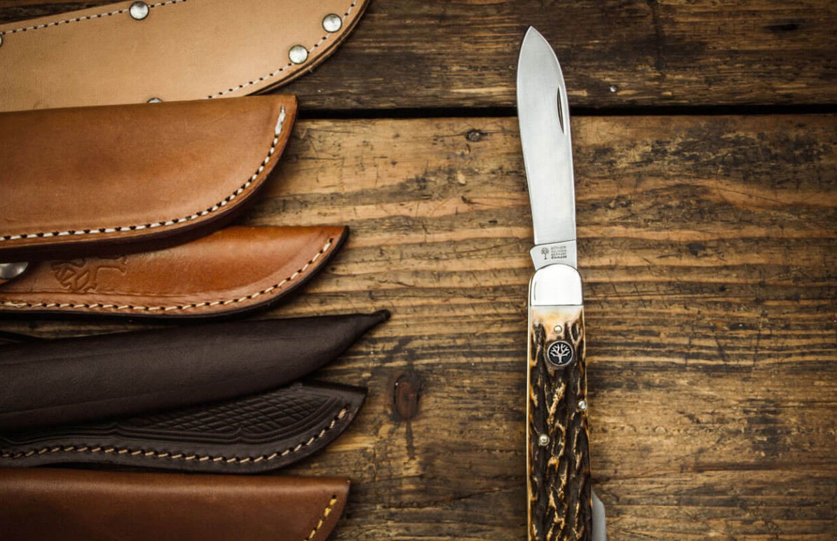 A stack of hand stitched leather sheaths beside a stag handled folding knife on a workbench